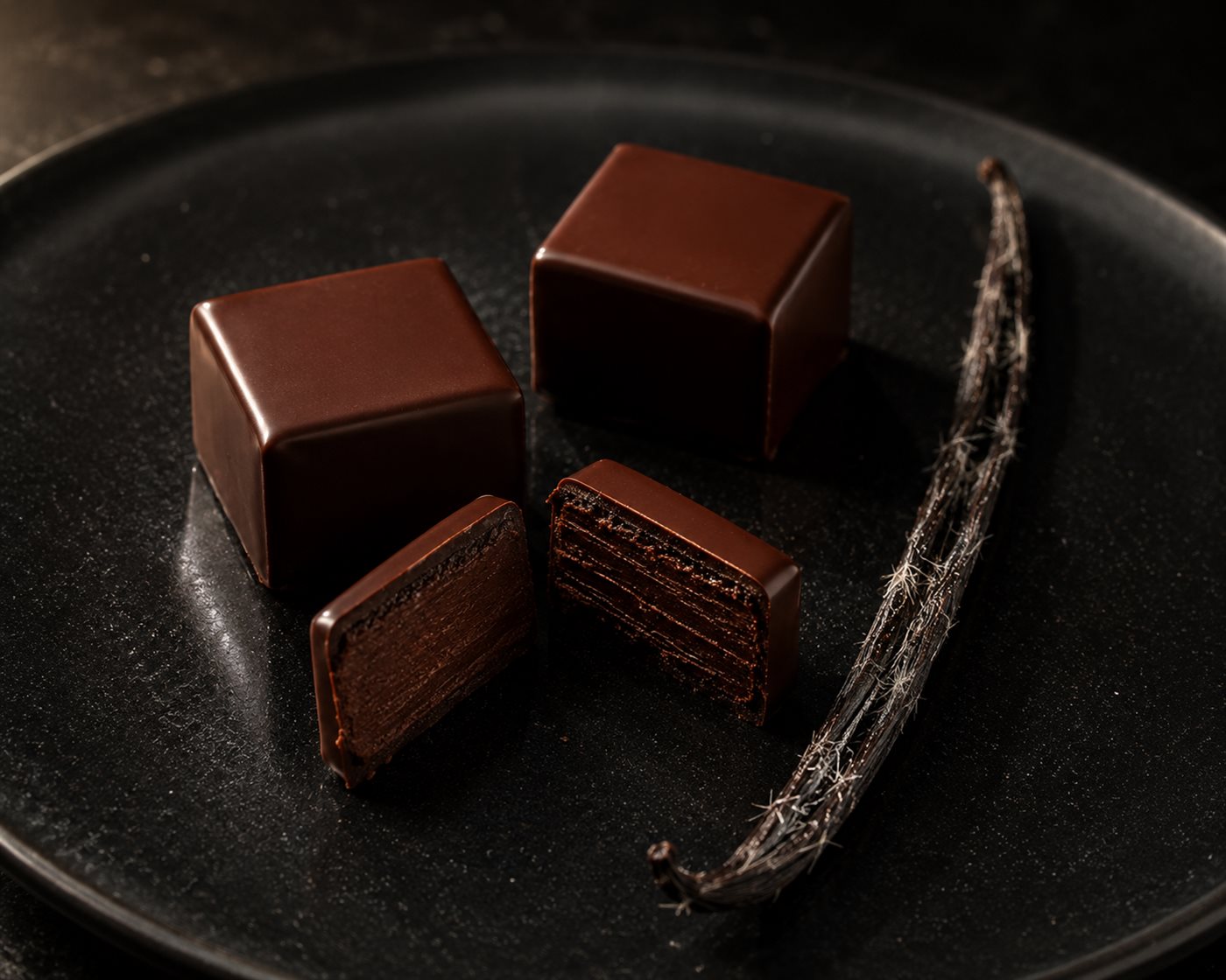 Bonbons de chocolat carrés et brillants, l'un coupé révélant la ganache, gousse de vanille givrée posée à côté