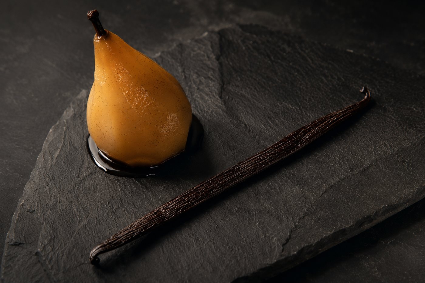 Poire pochée entière, brillante de sirop, posée sur une ardoise sombre à côté d'une gousse de vanille Pompona entière