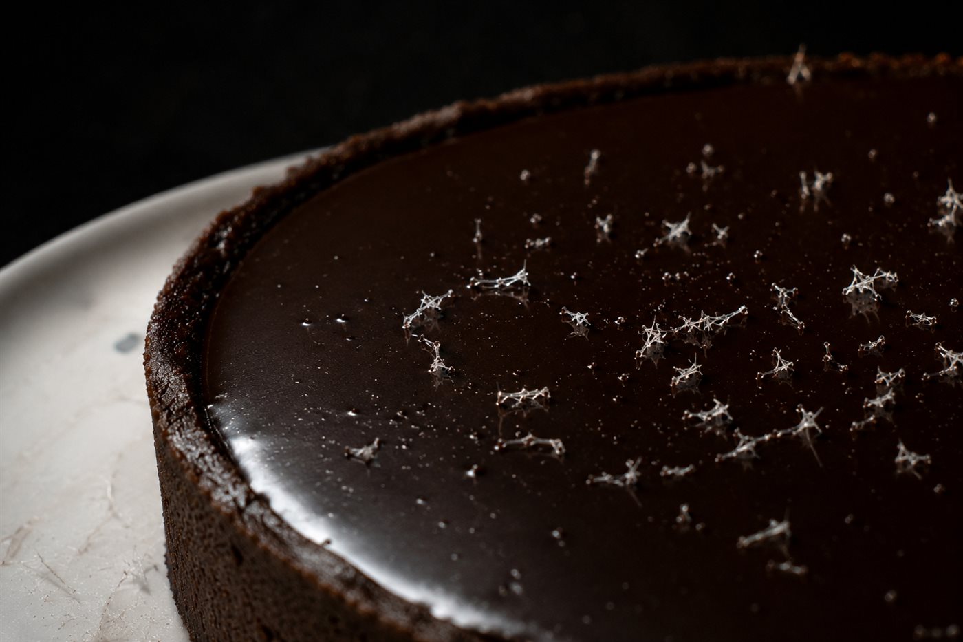 Tarte au chocolat noir lisse, cristaux de givre de vanille grattés en surface, contraste noir et blanc scintillant